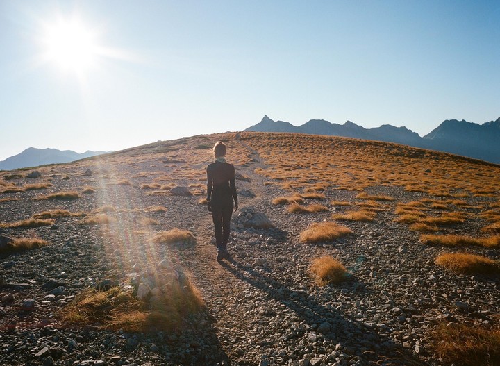 A woman walks on a rocky, grassy path toward mountains under a bright sun.