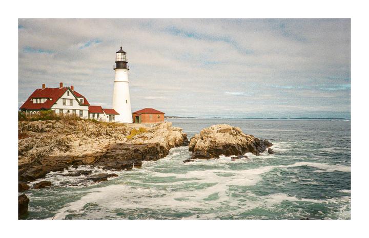 A white lighthouse and buildings with red roofs stand on a rocky coast with waves crashing against the shore.