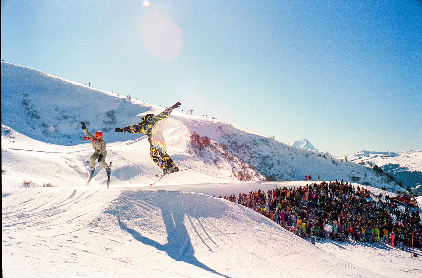 Two skiers are in mid-air performing tricks on a snowy mountain with a crowd of spectators watching.