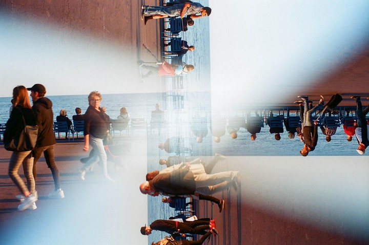 A multiple exposure shows people walking and sitting by the sea, with some figures appearing upside down and others right side up.