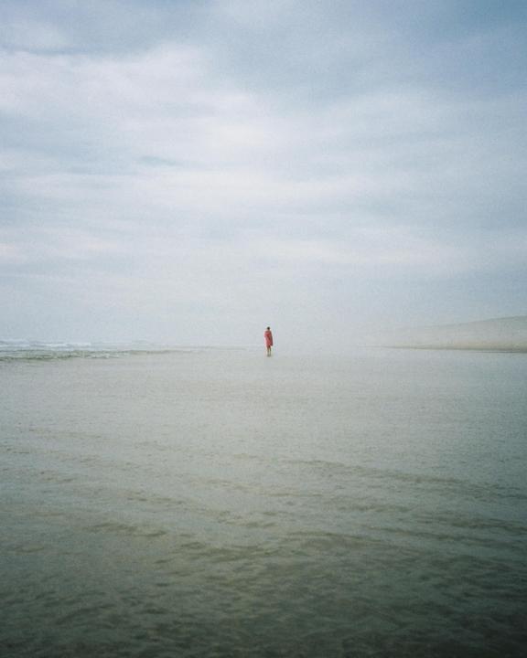 A person in a red coat stands in shallow water on a beach under a cloudy sky.