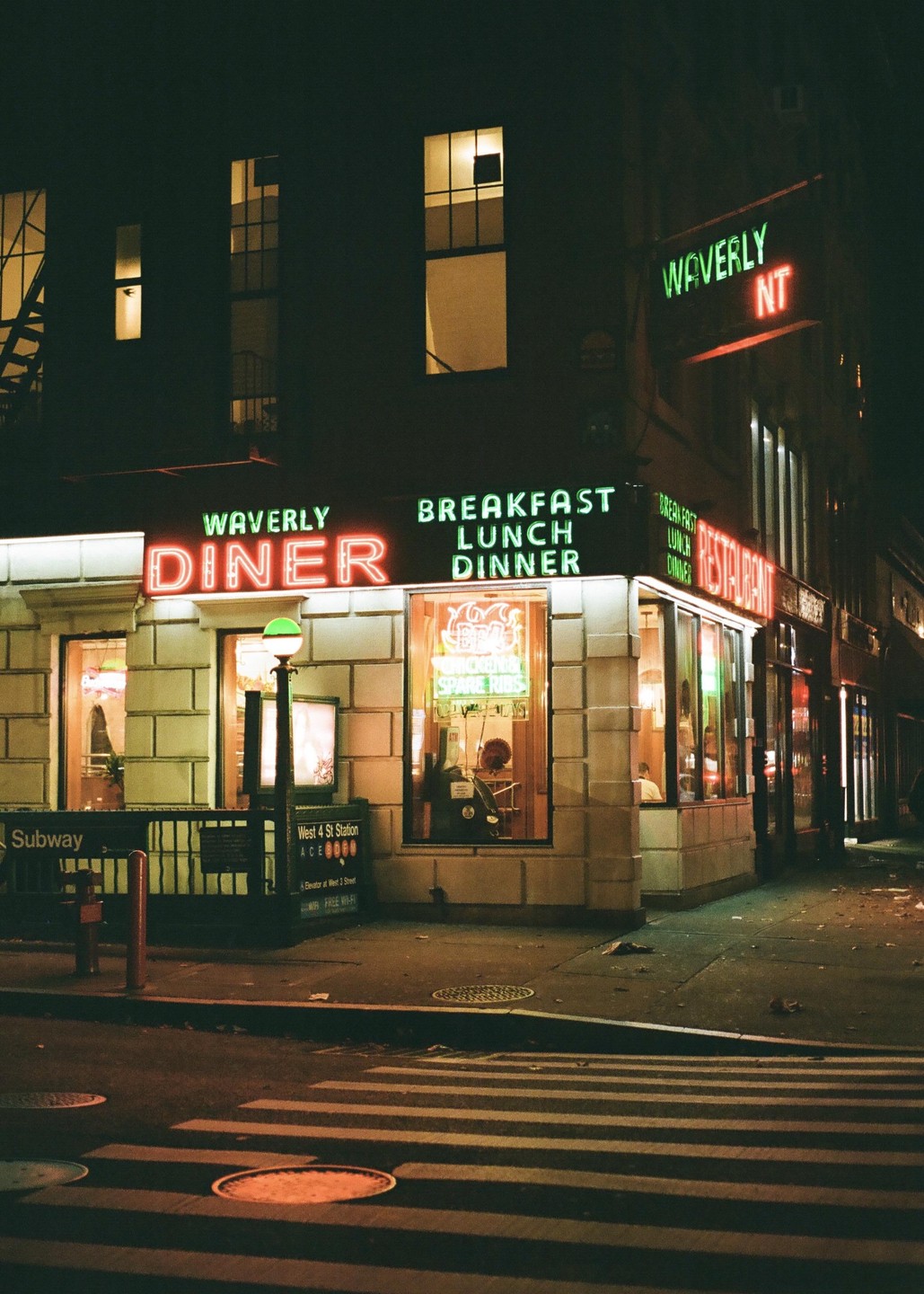 A diner with neon signs for breakfast, lunch, and dinner is visible at night next to a subway entrance.