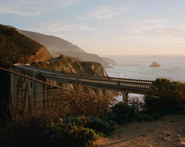 A bridge curves along a mountainous coastline overlooking the ocean at sunset.