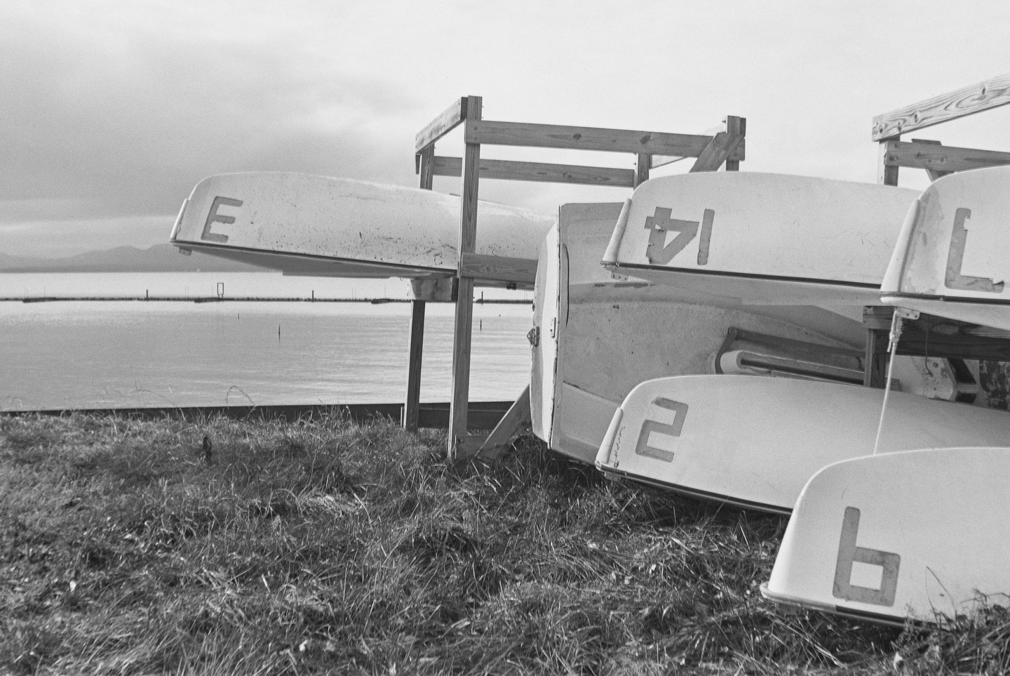 Several small boats are stored upside down on wooden racks next to a body of water with mountains in the distance.