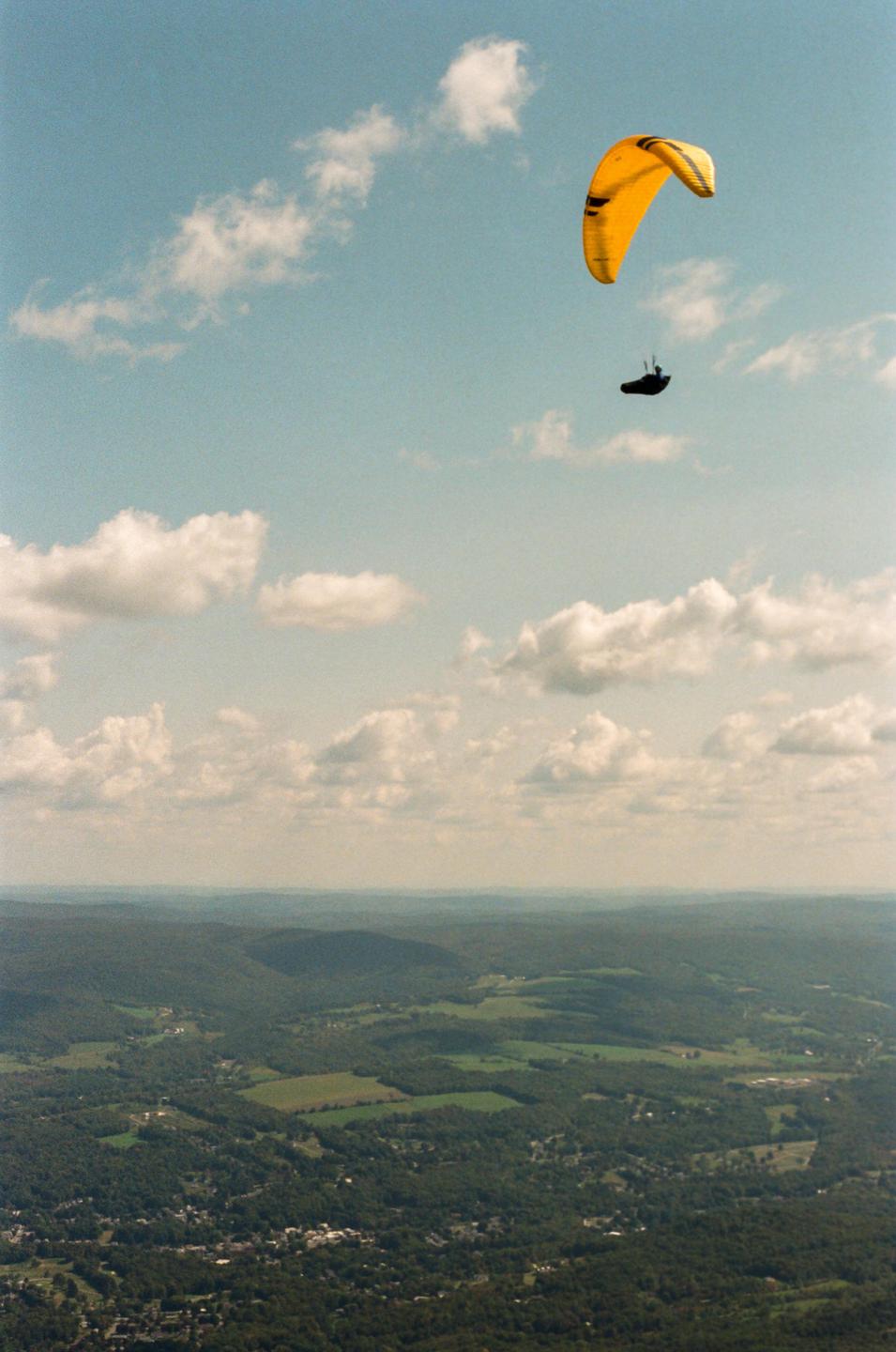 A paraglider with a yellow parachute flies high above a vast green landscape under a cloudy sky.