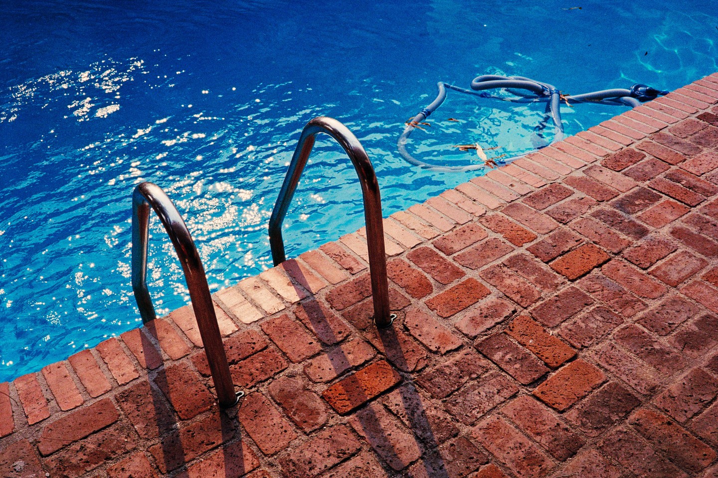 A swimming pool with a metal ladder and a brick patio is shown in bright sunlight.