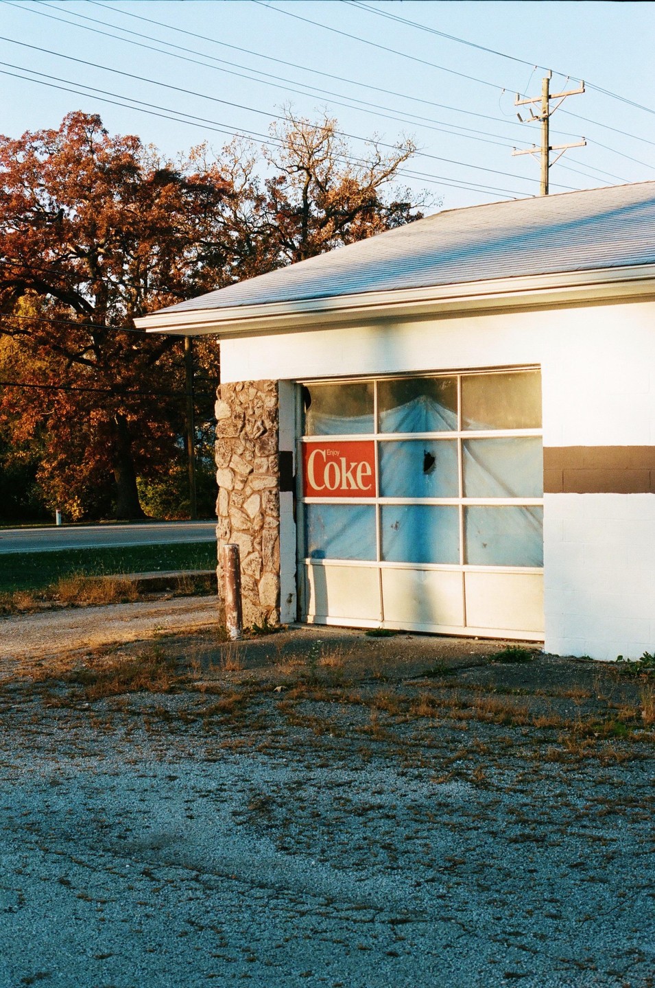 An old garage with a "Coke" sign on its window is bathed in the warm light of sunrise, with autumn trees in the background.