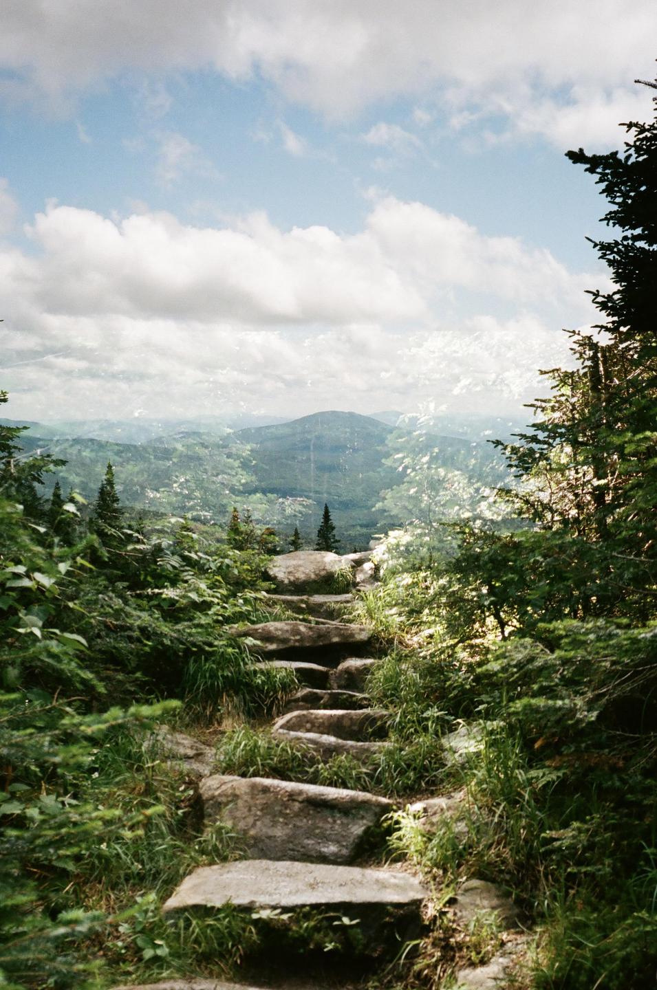 A rocky path leads up a mountain with a view of distant mountains and a cloudy sky.