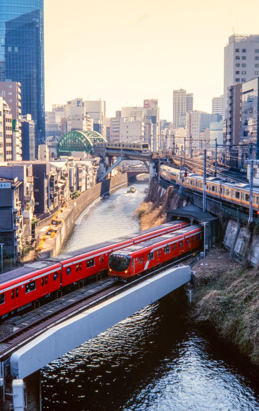 Two red trains are on tracks above a canal with a small boat, surrounded by buildings and other train lines.