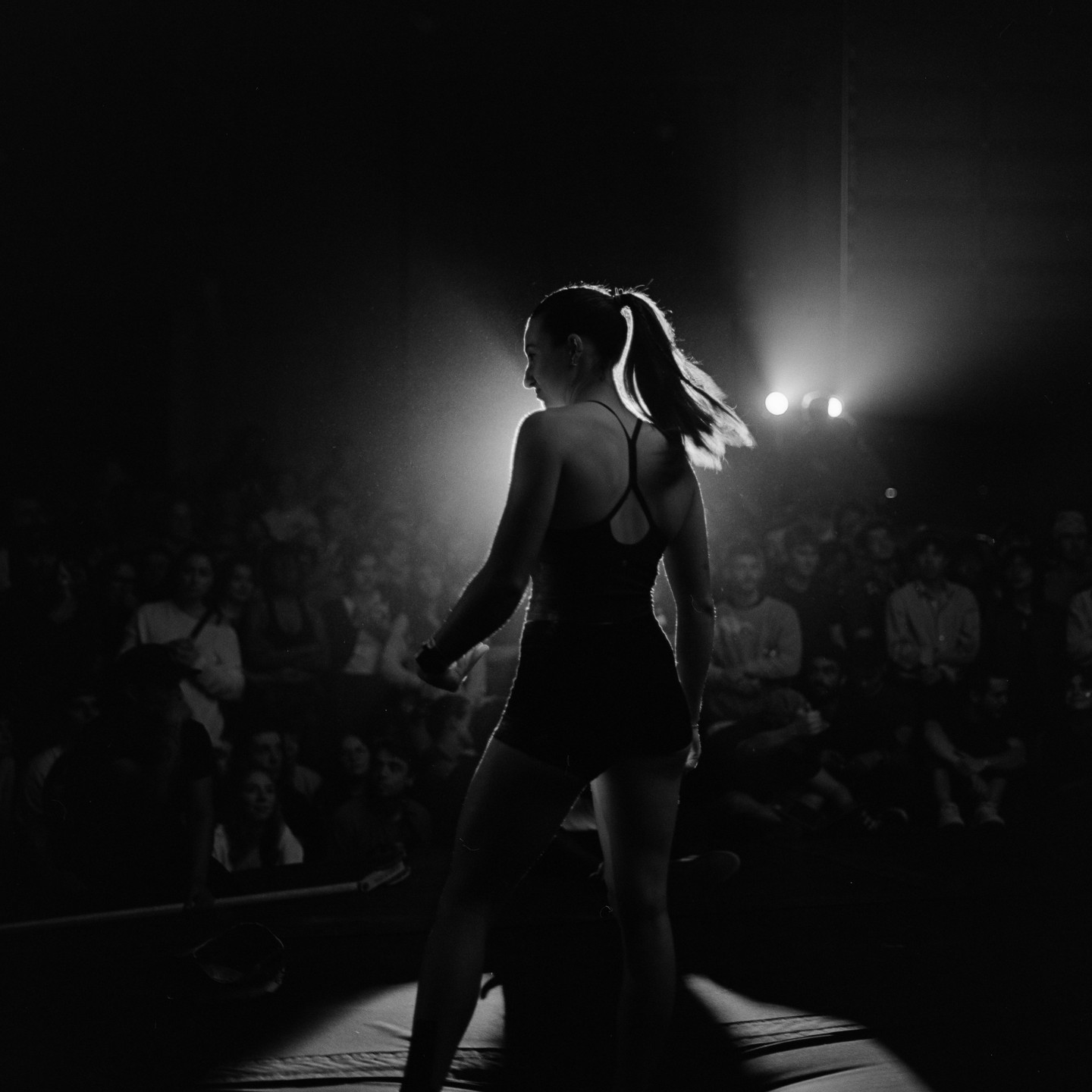 A woman stands on a stage, backlit by a spotlight, facing a crowd in a dark room.