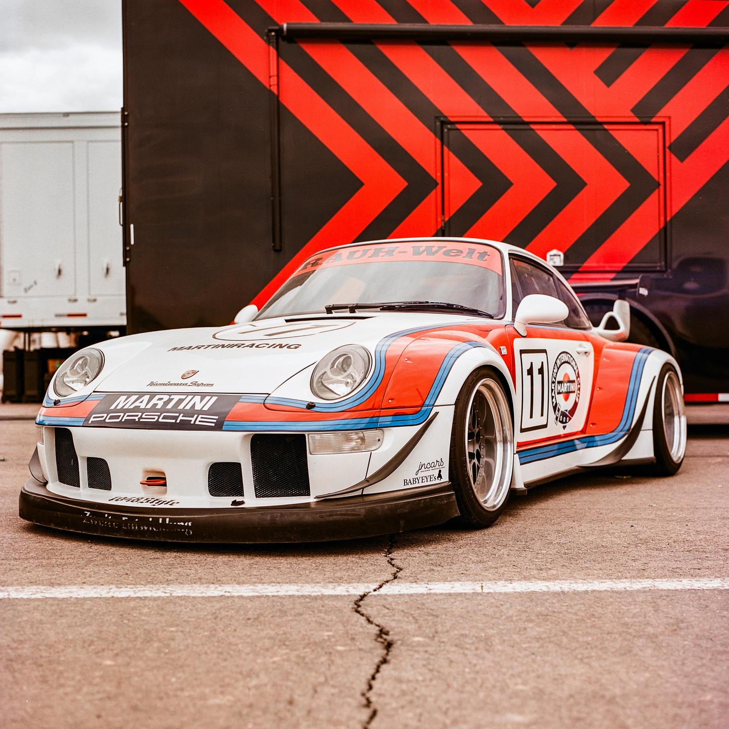 A white Porsche race car with red and blue stripes is parked on asphalt in front of a black and red striped trailer.