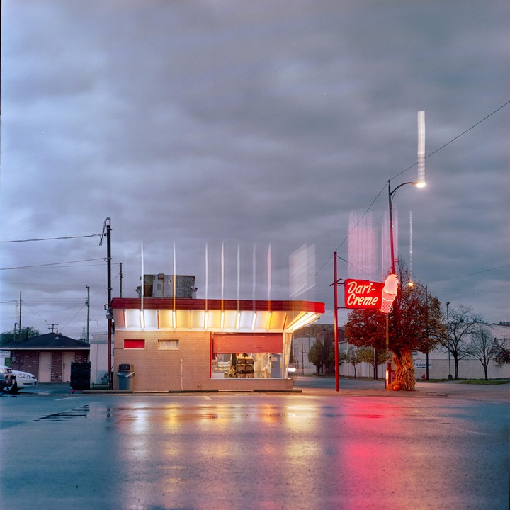 A Dari-Creme ice cream stand with a red neon sign is illuminated at dusk, reflecting on the wet pavement.
