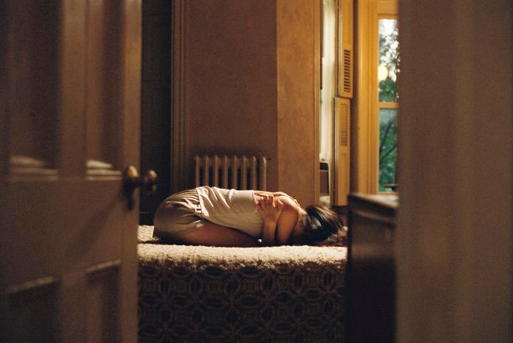 A woman lies curled on a bed in a dimly lit room, with light coming from a window on the right.