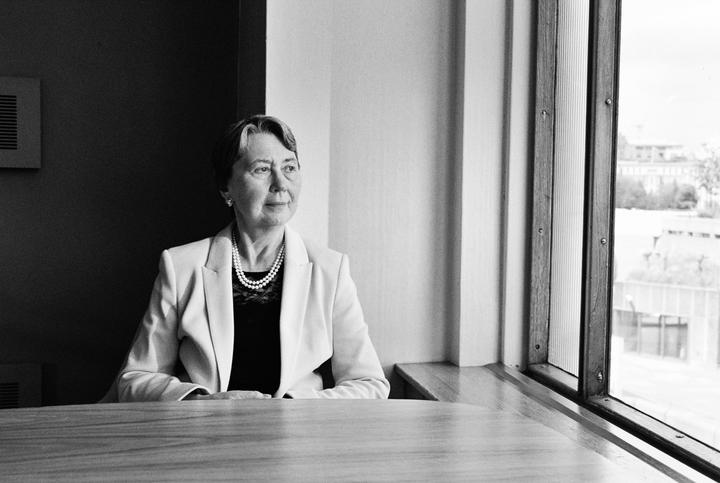A woman wearing a blazer and pearl necklace sits at a table looking out a window.