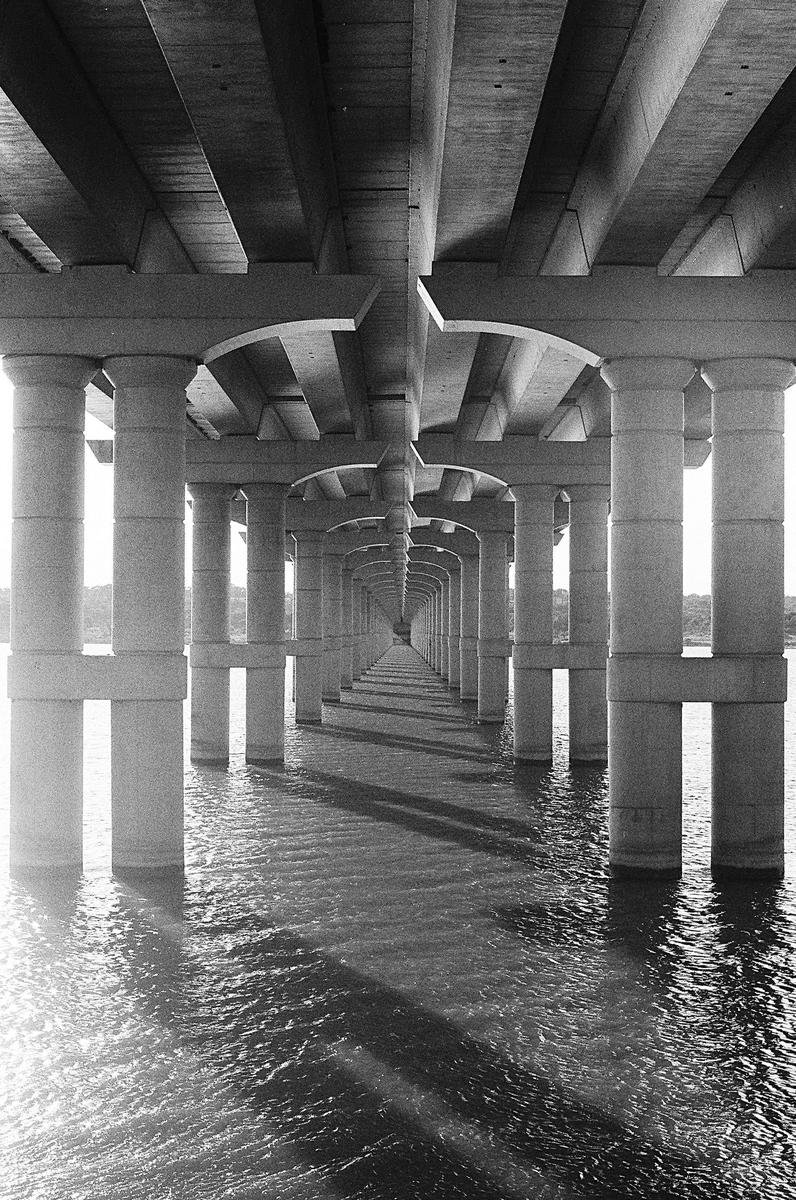 The concrete pillars of a bridge extend into the distance over rippling water, creating a tunnel-like effect.