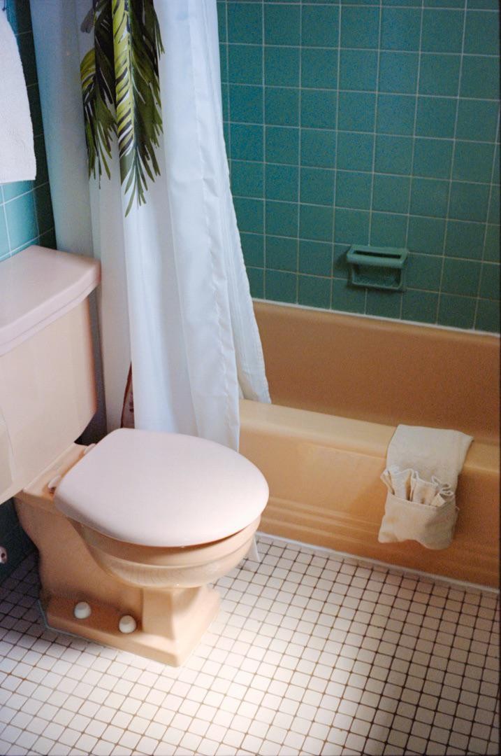A peach toilet and bathtub are next to a white shower curtain with a green palm leaf pattern in a bathroom with teal wall tiles and white floor tiles.