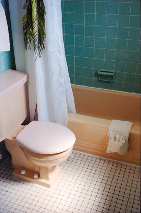 A peach toilet and bathtub are next to a white shower curtain with a green palm leaf pattern in a bathroom with teal wall tiles and white floor tiles.