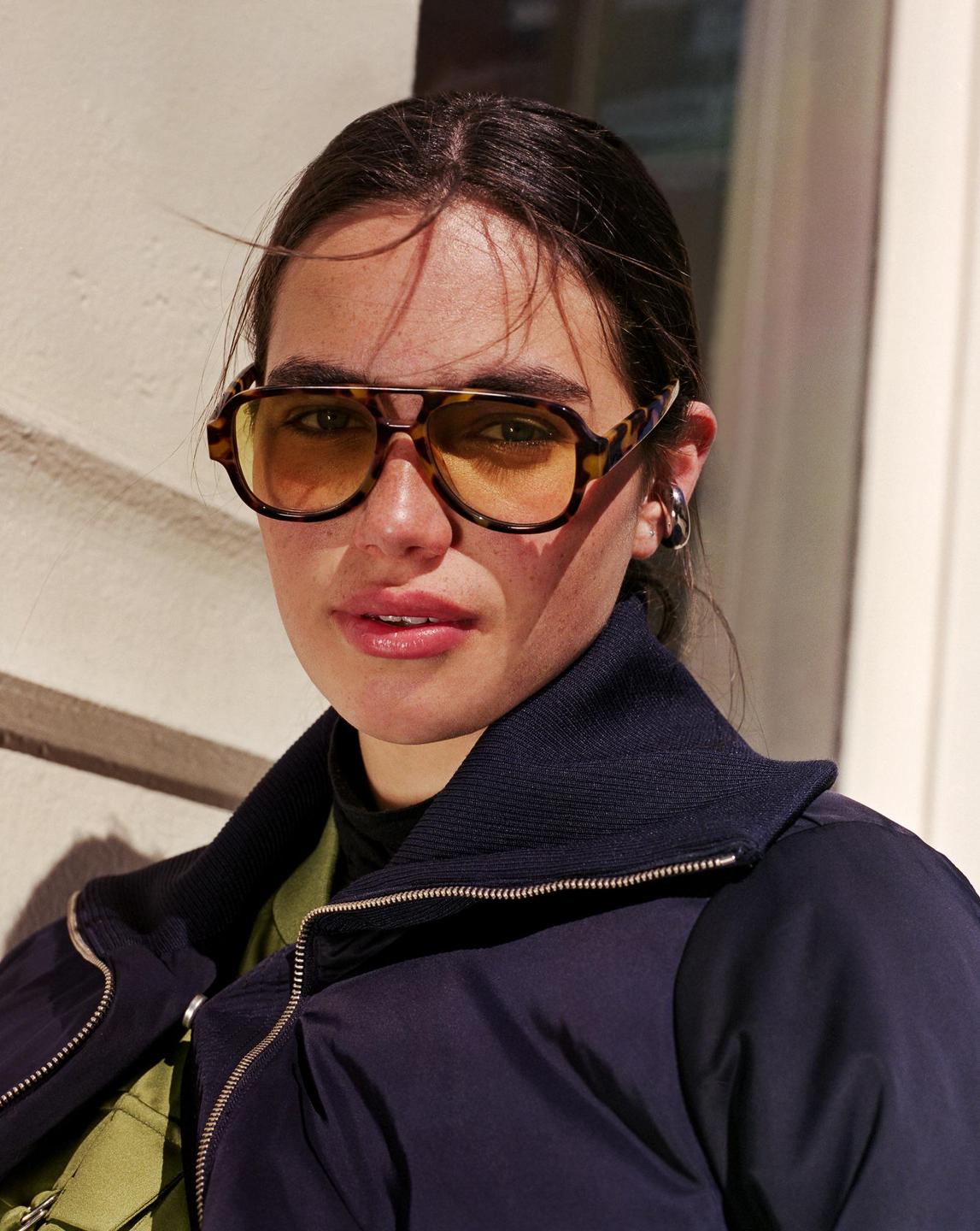 A woman wearing sunglasses and a jacket looks directly at the viewer.