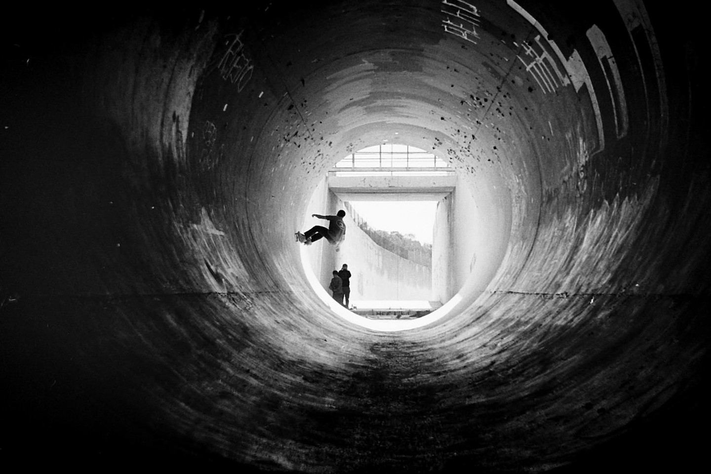 A skateboarder rides a full pipe while two people watch from the end of the tunnel.