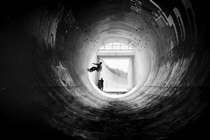 A skateboarder rides a full pipe while two people watch from the end of the tunnel.
