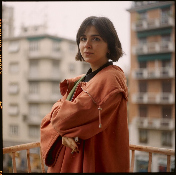 A woman in an orange jacket stands on a balcony with buildings in the background.