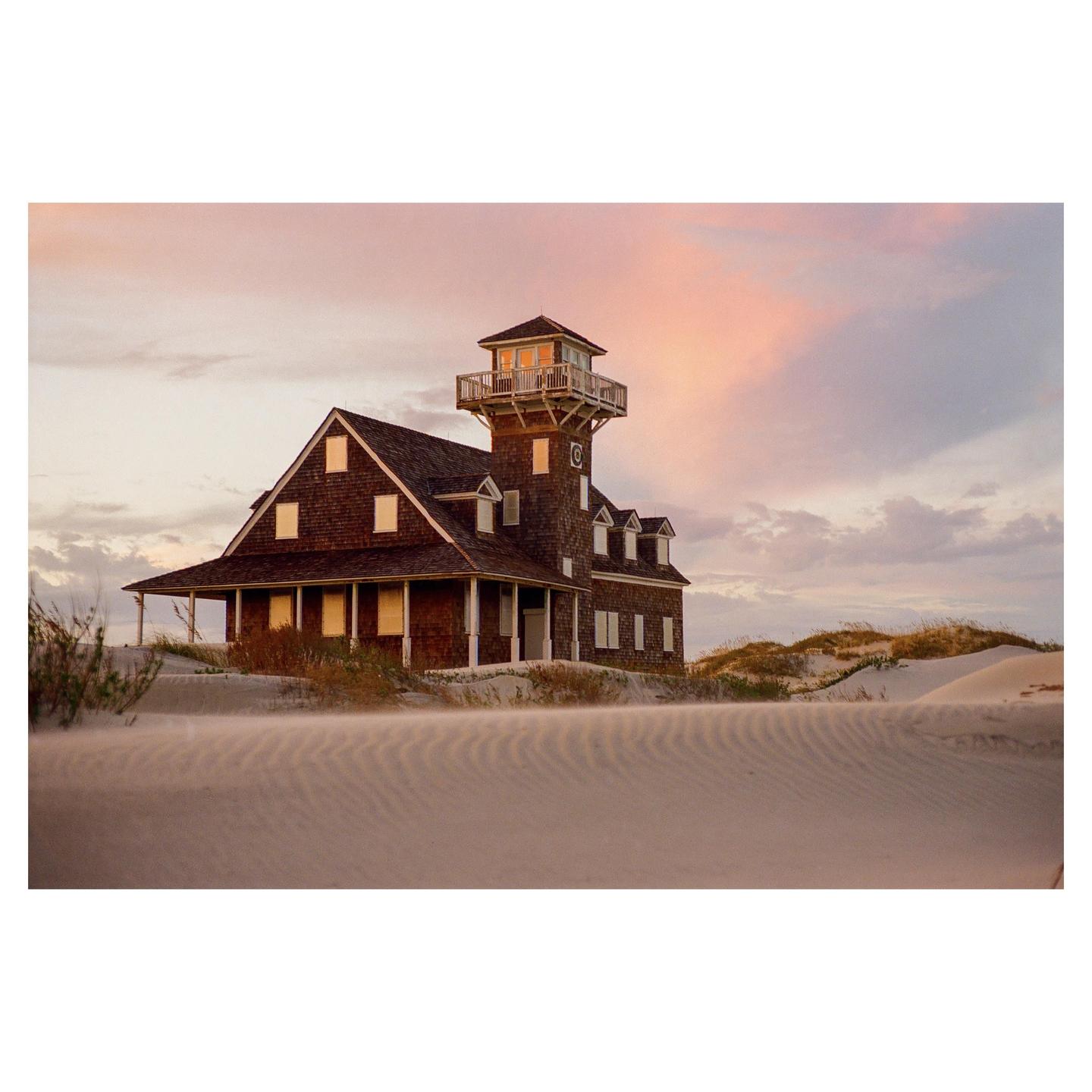 A wooden house with a tower stands on a sandy dune under a pink and purple sky.