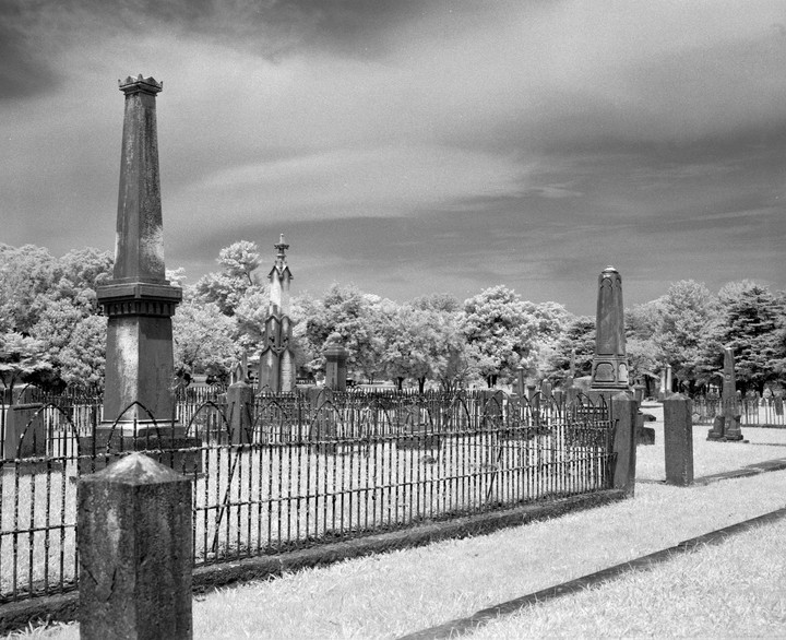A black and white photograph shows a cemetery with many tombstones and a metal fence in the foreground.