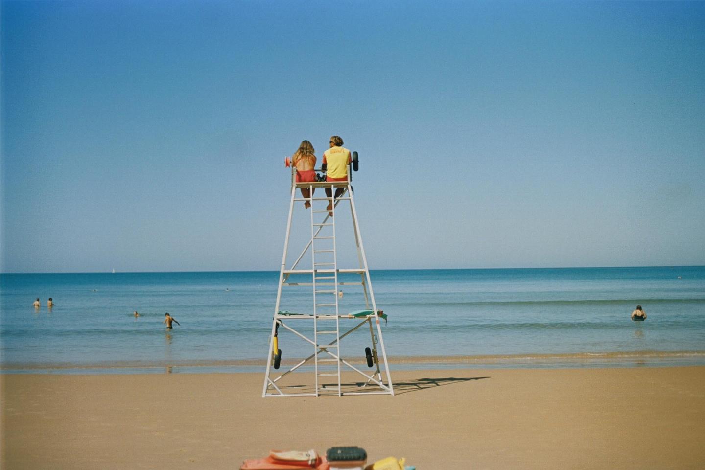 A couple sits on a lifeguard stand overlooking a beach with people swimming in the ocean.