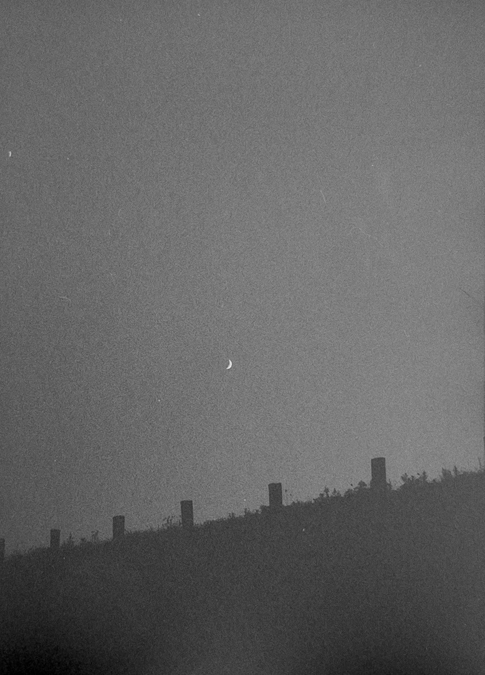 A crescent moon hangs in a hazy sky above a dark hill with fence posts.