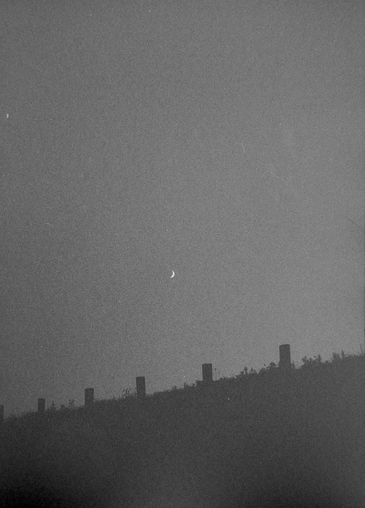 A crescent moon hangs in a hazy sky above a dark hill with fence posts.
