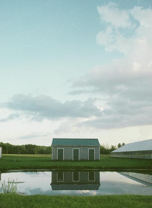 A small building with a green roof is reflected in a pond under a cloudy sky.