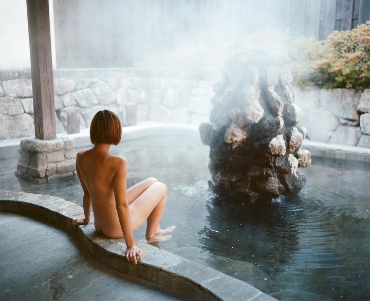 A woman sits on the edge of a steaming outdoor bath with a rock formation in the water.