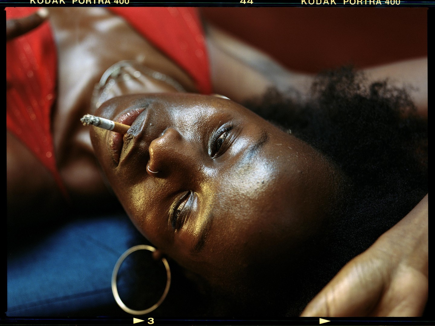 A woman with a cigarette in her mouth is lying down, looking up, with a hoop earring visible.