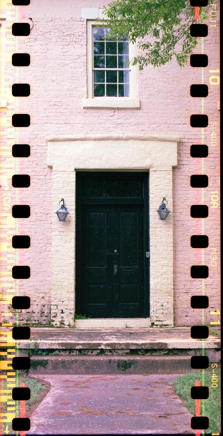 A dark green door with two lanterns on either side is set into a light pink brick wall, with a window above the door and a stone path leading up to it.