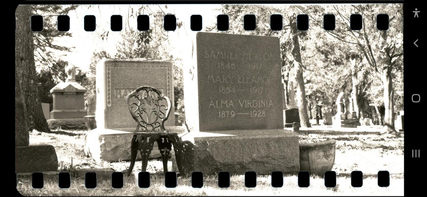 A black and white photo shows a cemetery with a decorative metal chair in front of a large tombstone.