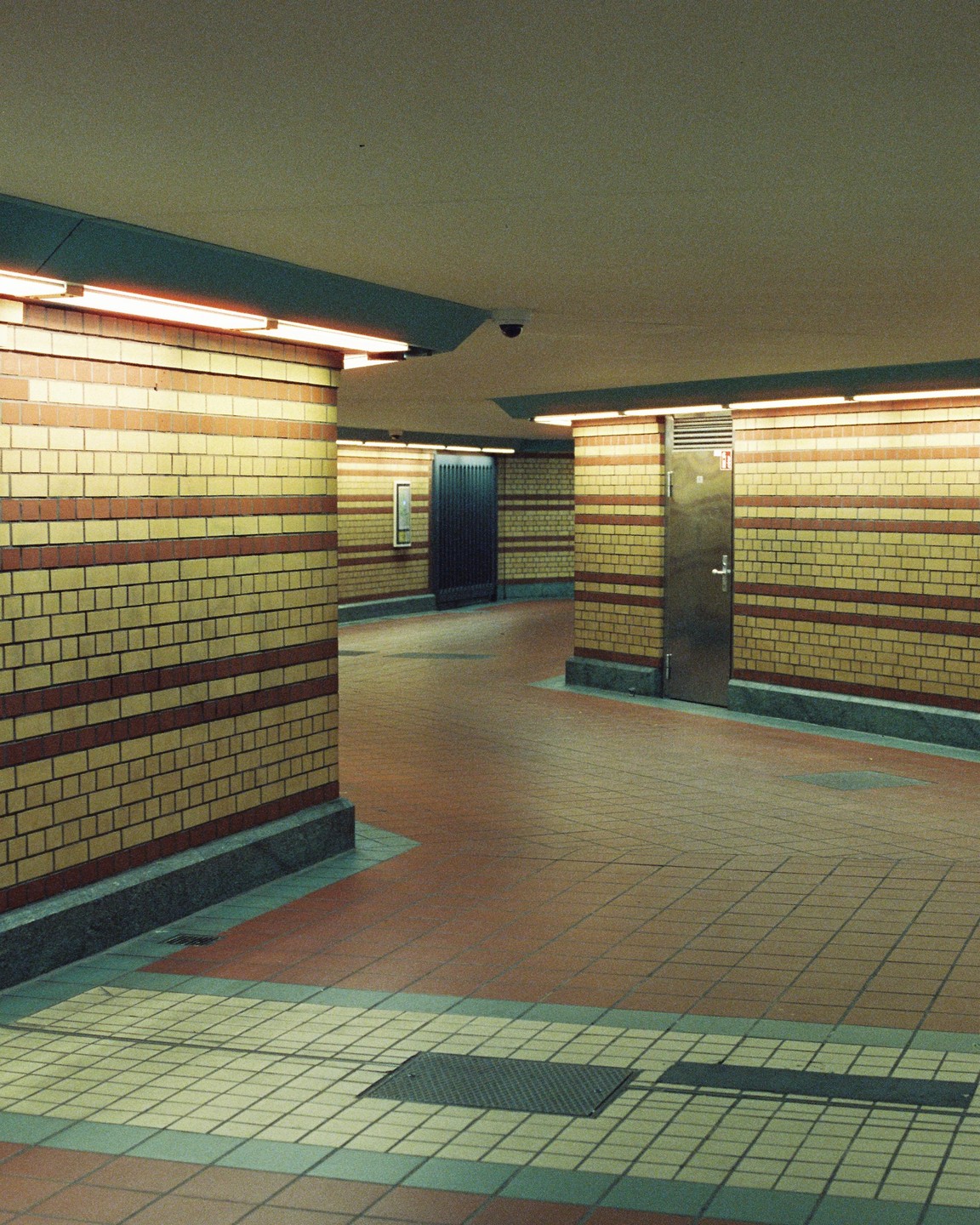 An empty underground walkway is illuminated by fluorescent lights, revealing tiled walls and floors.