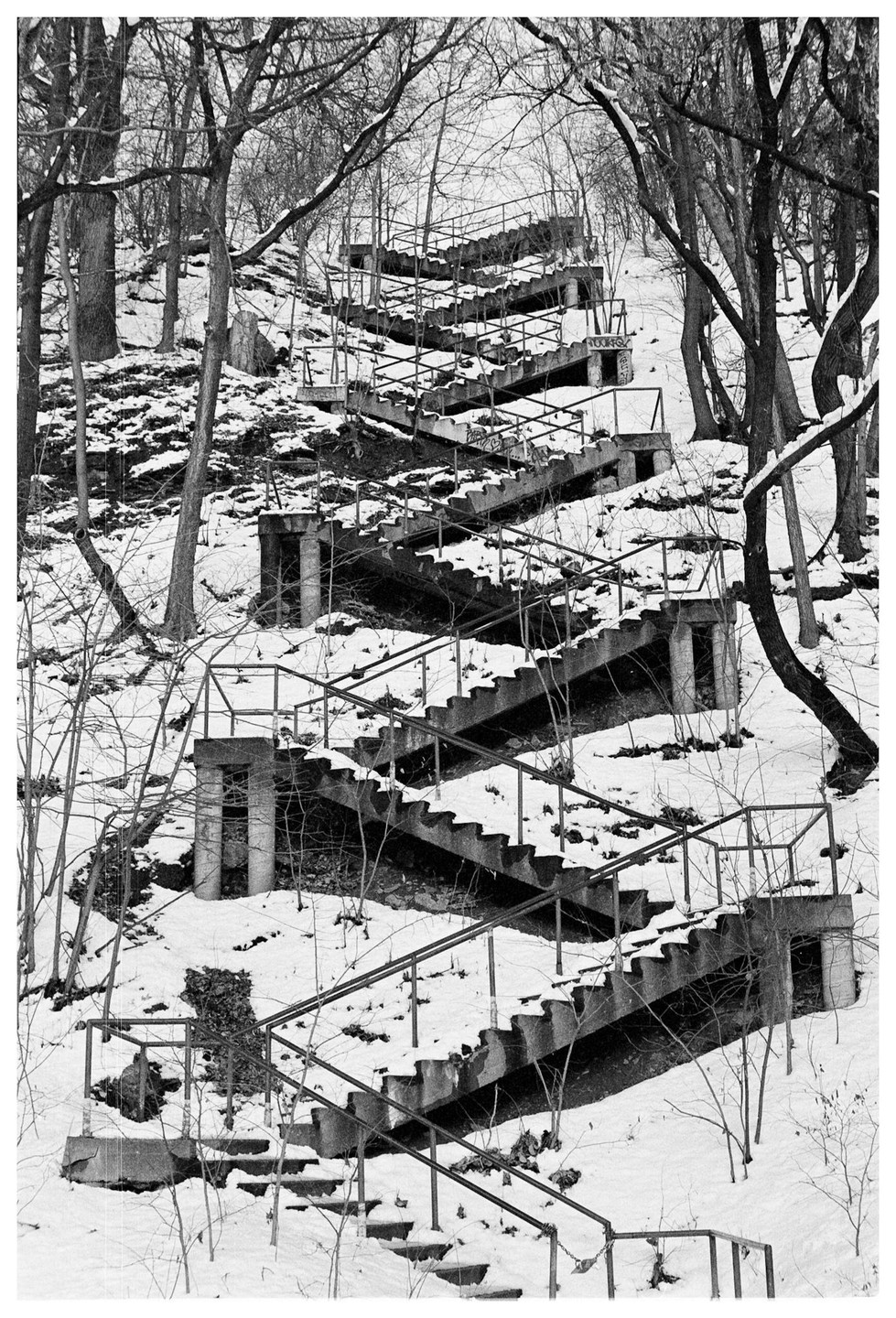 A long, winding concrete staircase with metal railings ascends a snow-covered hill surrounded by bare trees.