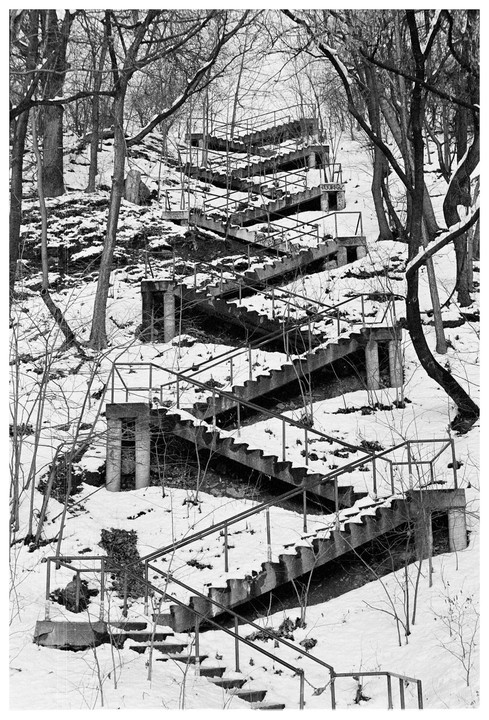 A long, winding concrete staircase with metal railings ascends a snow-covered hill surrounded by bare trees.