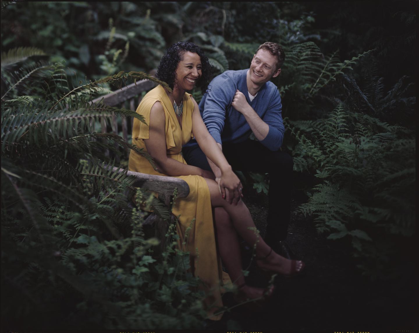 A couple sits on a wooden bench surrounded by ferns.