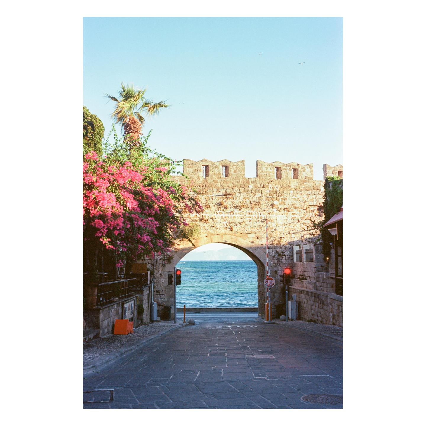 A stone archway frames a view of the Mediterranean Sea, with pink bougainvillea and a palm tree on the left.