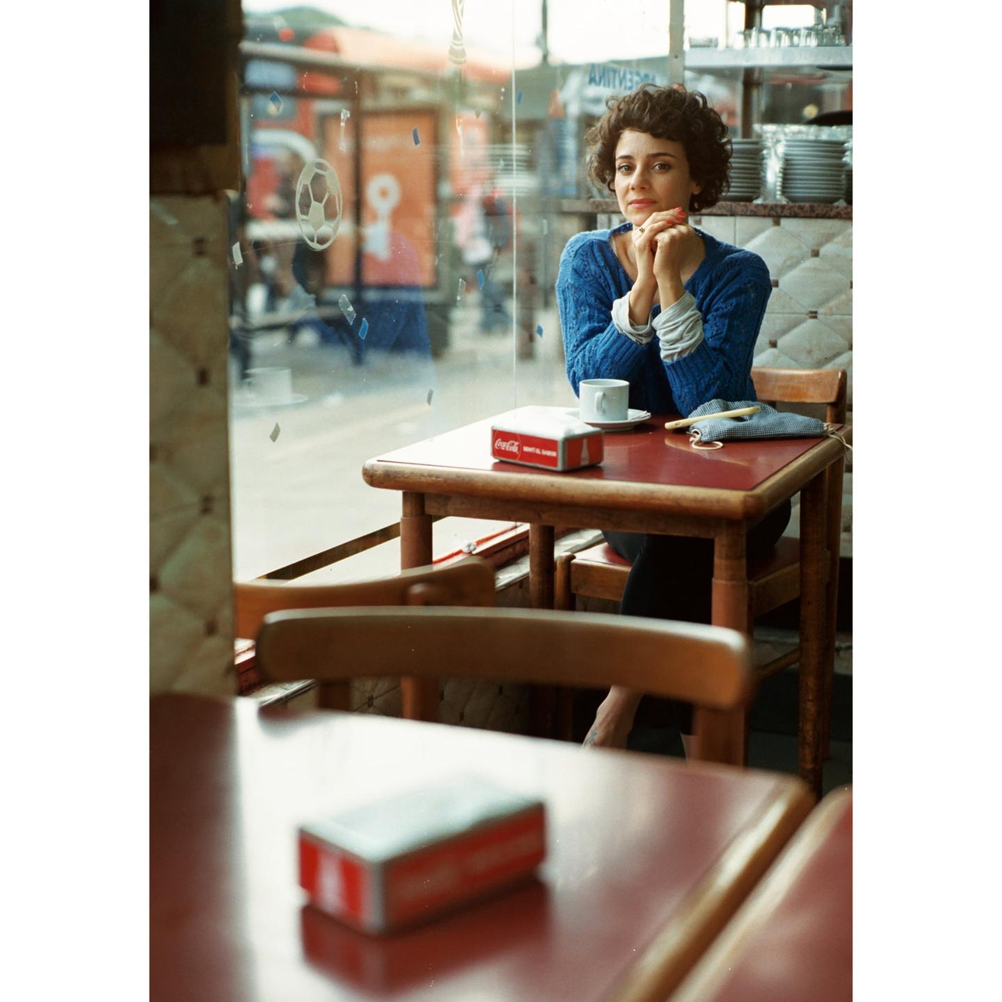 A woman with curly hair sits at a cafe table with a cup and a Coca-Cola napkin dispenser.