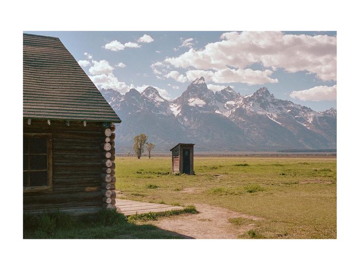 A wooden cabin and an outhouse stand in a grassy field with snow-capped mountains in the background under a cloudy sky.