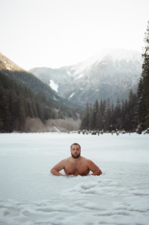 A man is chest-deep in a frozen lake surrounded by snowy mountains and pine trees.
