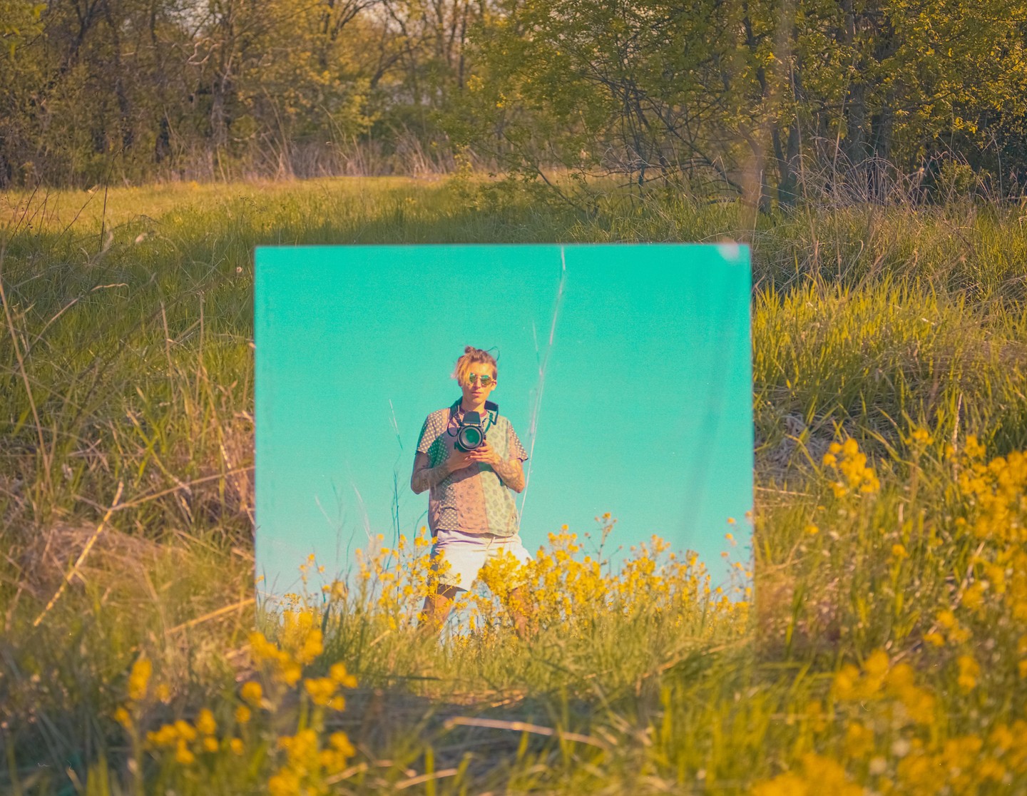 A person holding a camera is reflected in a mirror placed in a field of yellow flowers and green grass.