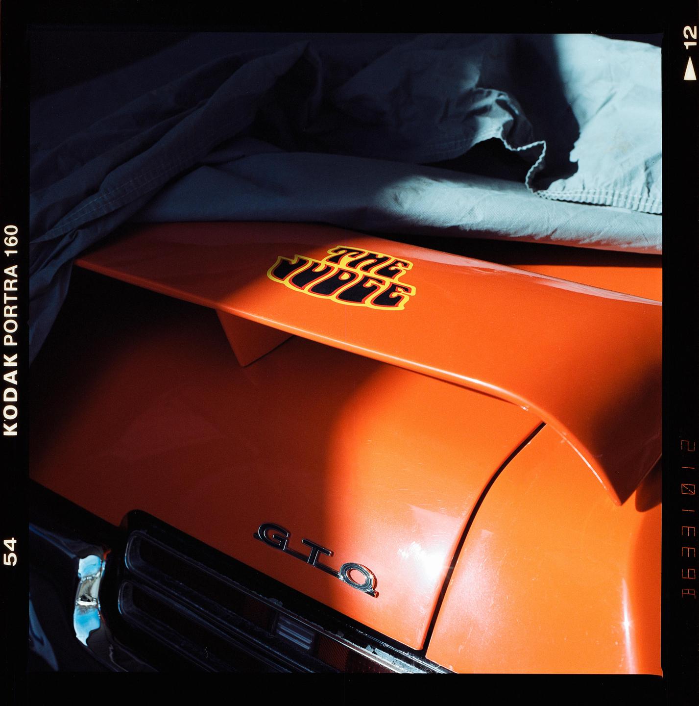The rear of an orange car with a spoiler and a "The Judge" decal is partially covered by a gray sheet.