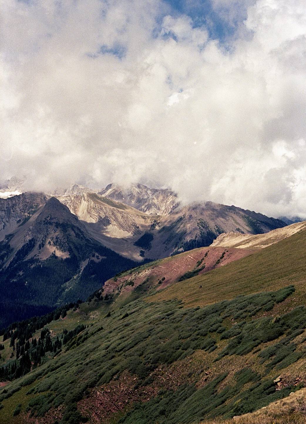Clouds hang over a mountain range with a forested valley and a grassy slope in the foreground.