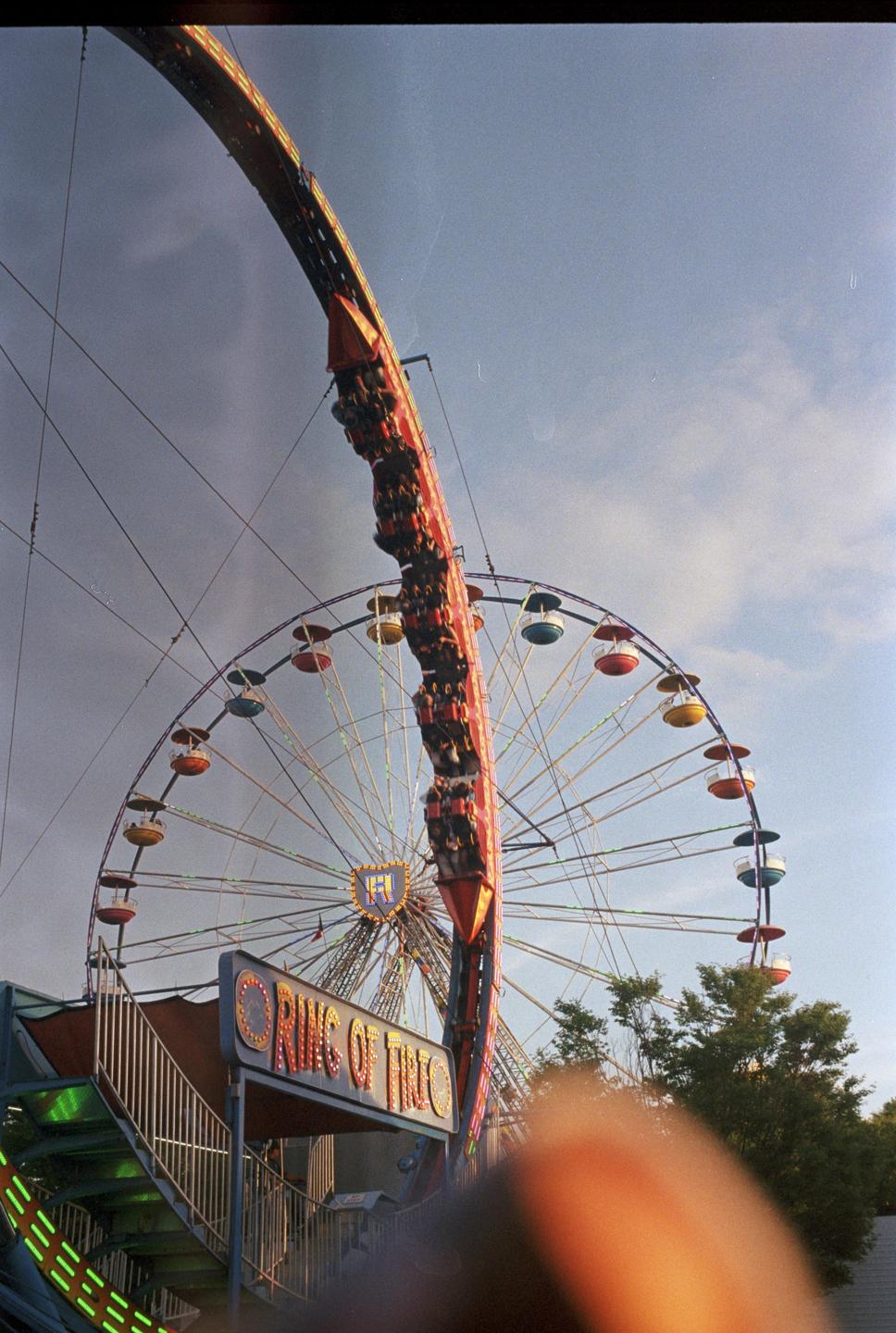 A large amusement park ride with people on it is visible against a cloudy sky, with a ferris wheel in the background.