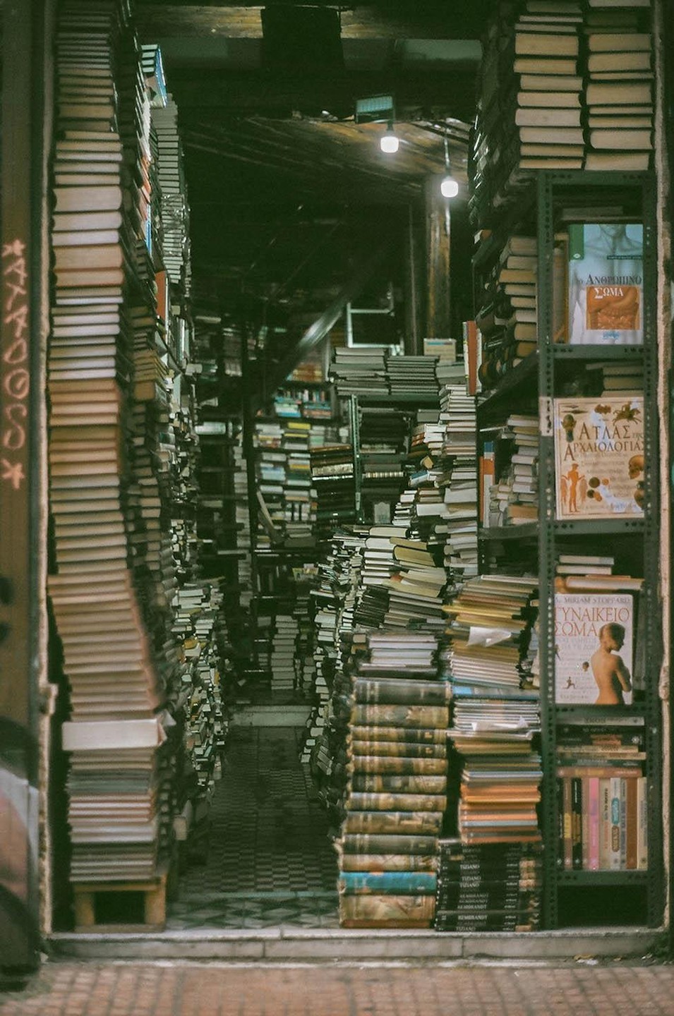 A narrow aisle in a bookstore is filled with stacks of books from floor to ceiling.