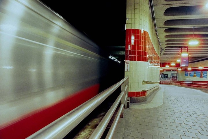 A subway train blurs past a tiled station platform with red and white walls.