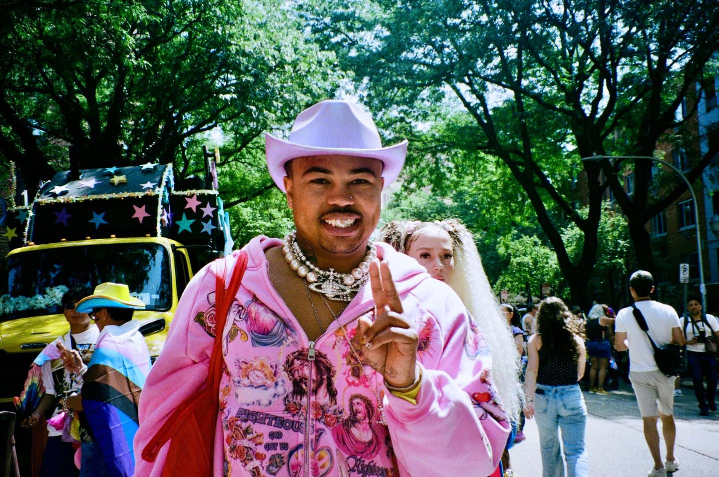 A person in a pink hoodie and a lavender cowboy hat smiles at the camera, holding up two fingers.